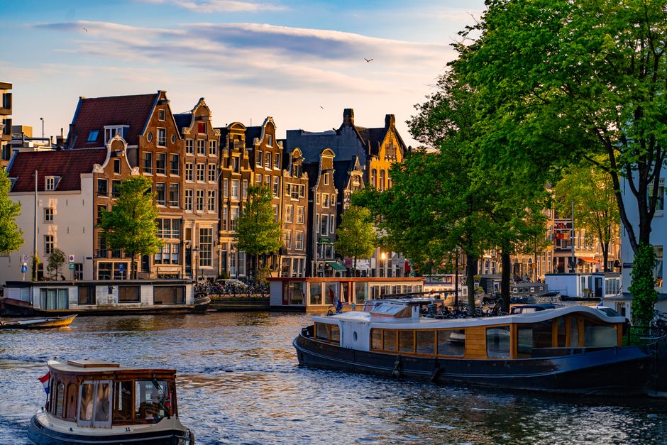 Scenic view of Amsterdam canal houses and boats at sunset with vibrant green trees.