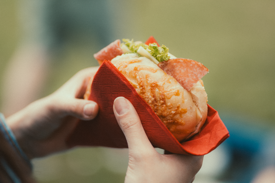 A close-up of hands holding a sandwich with salami and vegetables in an outdoor setting.