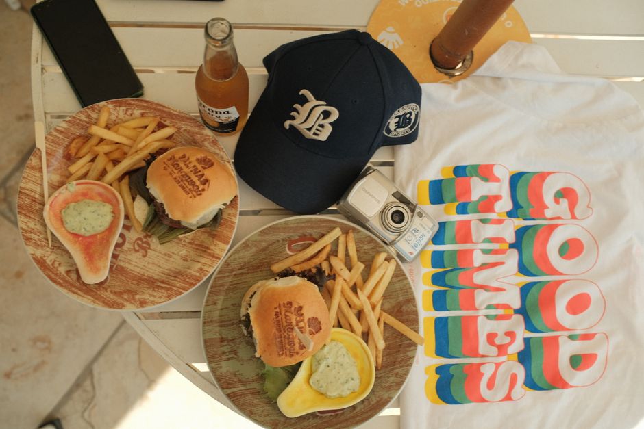Flat lay of burgers with fries, cap, and camera on a table setting emphasizing good times.