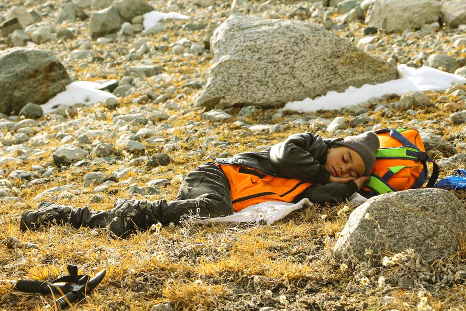 A young trekker rests on a rocky mountainside in Khyber Pakhtunkhwa, Pakistan, surrounded by snow-dusted boulders.