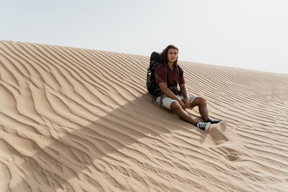 A young man in casual wear sitting on a desert sand dune, enjoying the tranquil landscape.