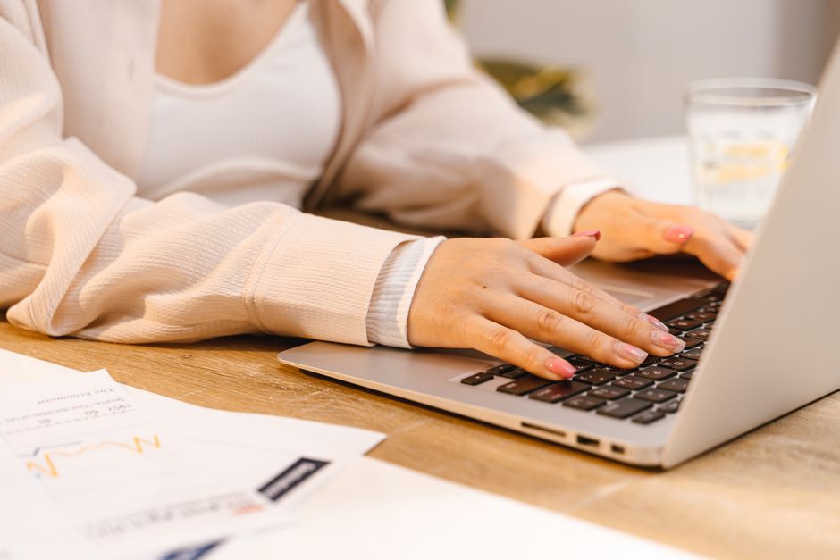Close-up of a woman's hands typing on a laptop with documents on a wooden desk.