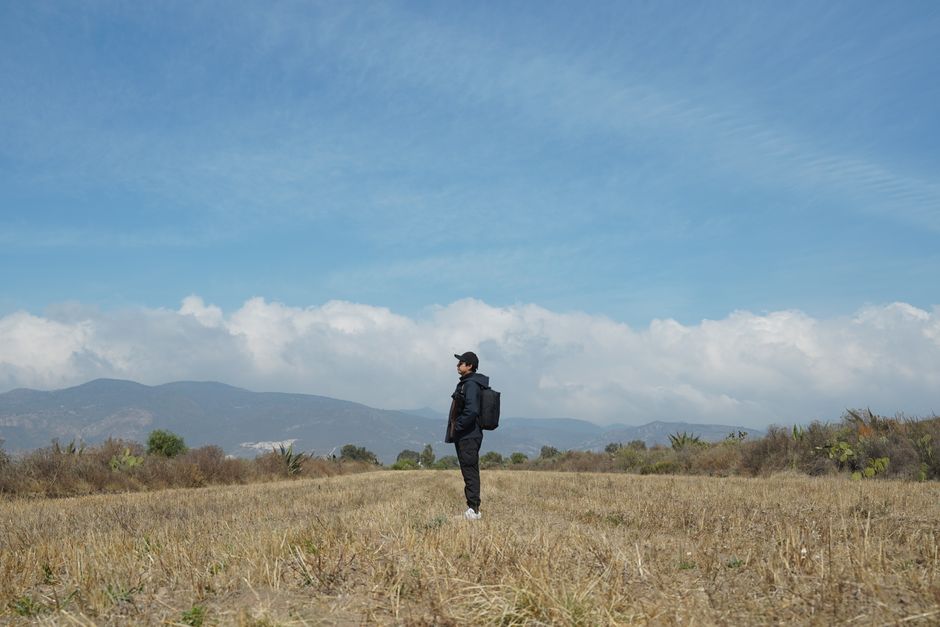 A lone traveler stands in an open field under a vast sky, surrounded by mountain scenery.