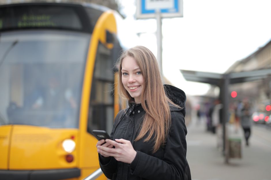 Smiling woman texting on phone at tram station in city street setting.