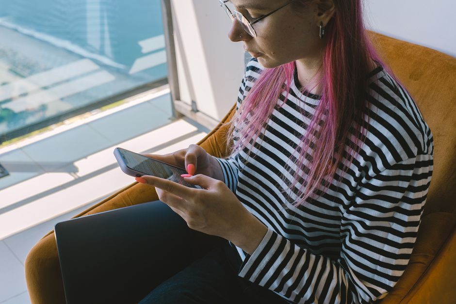 A young woman with pink hair using a smartphone, seated by a window with city view.