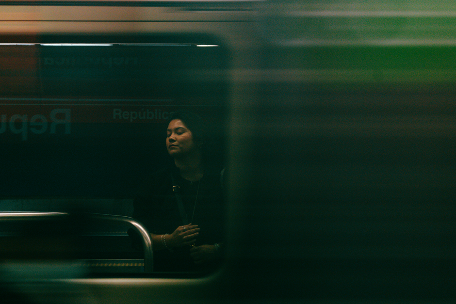 A woman stands on a subway platform with a passing train, creating a motion blur effect.