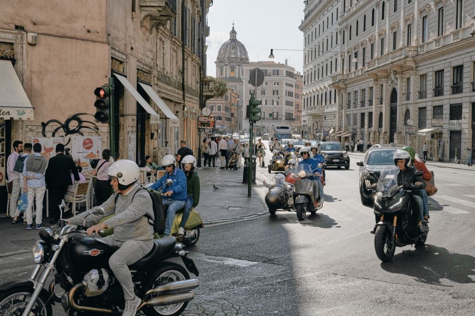 Daytime scene of scooters and pedestrians on a bustling street in Rome, Italy, with classic architecture.