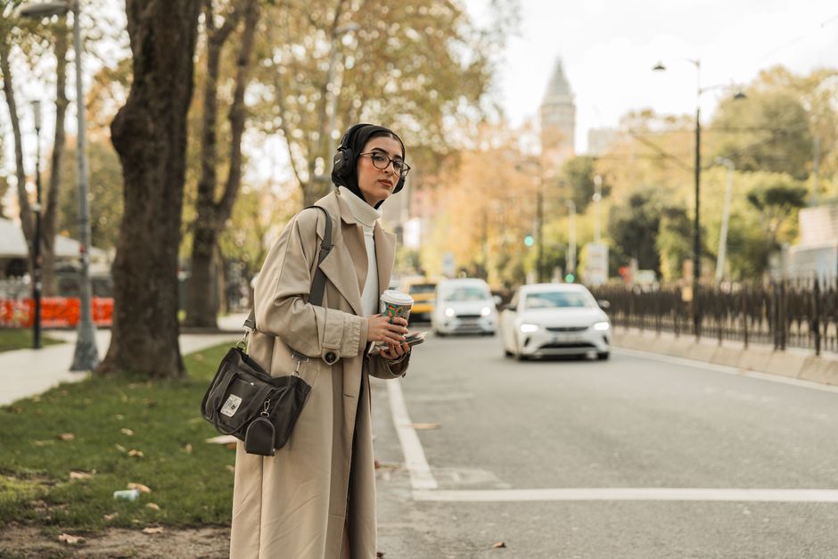 Woman in coat holding coffee, listening with headphones on city street during autumn.