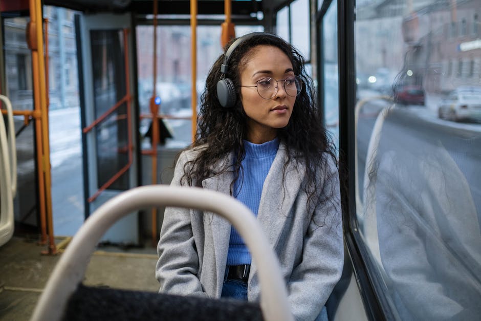 Young woman in glasses and headphones sitting on a city bus, looking out the window during the day.