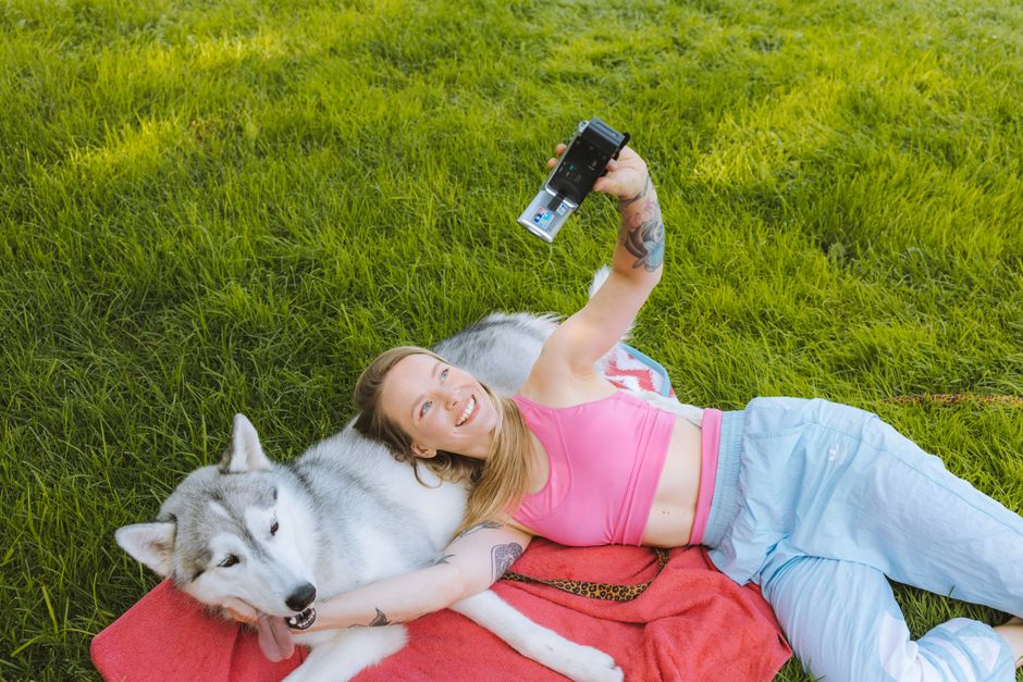 A cheerful woman in a pink tank top takes a selfie with her Siberian Husky while lying on a red blanket in a grassy park.