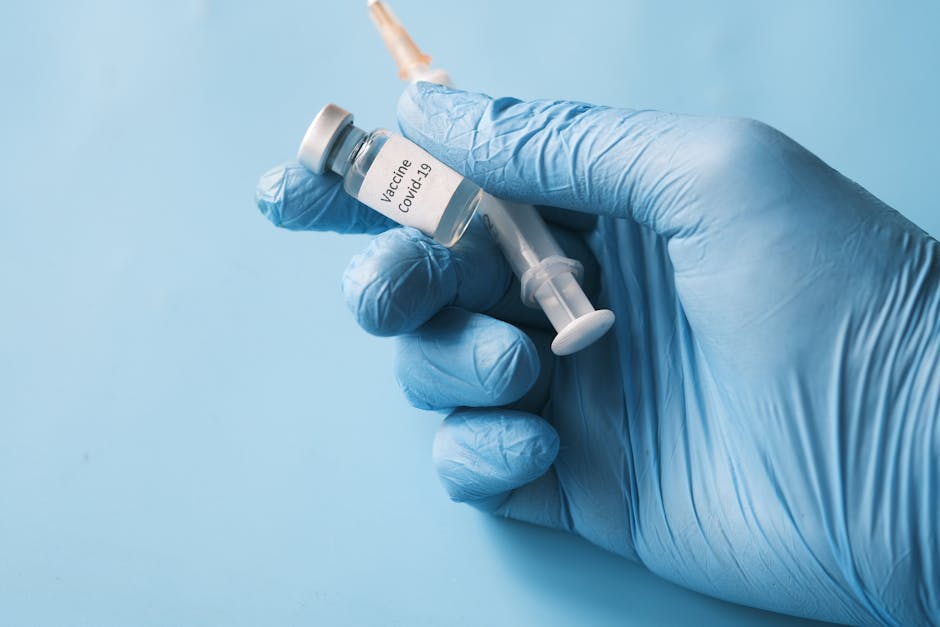 Blue gloved hand holding a COVID-19 vaccine vial and syringe on a blue background.
