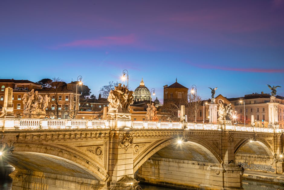 Illuminated Sant'Angelo Bridge with St. Peter's Basilica in Rome at twilight, showcasing historic architecture.